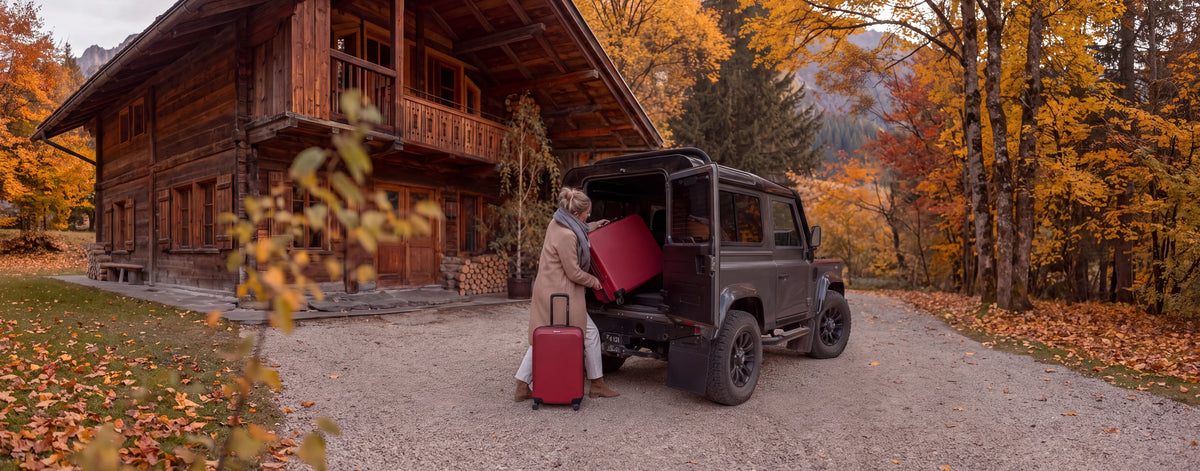 Set de valises Travel World rouge chargé dans un Land Rover Defender devant un chalet de montagne en automne